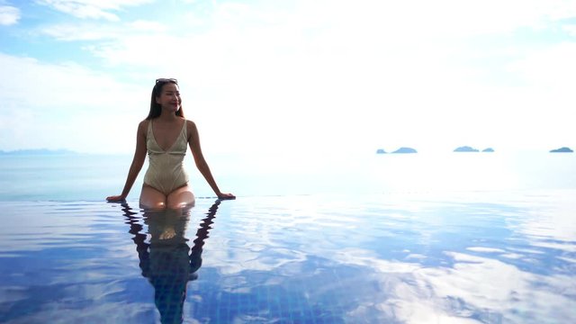 Beautiful asian woman sitting by the infinity pool, sea background. Holiday scene with copy space for text.