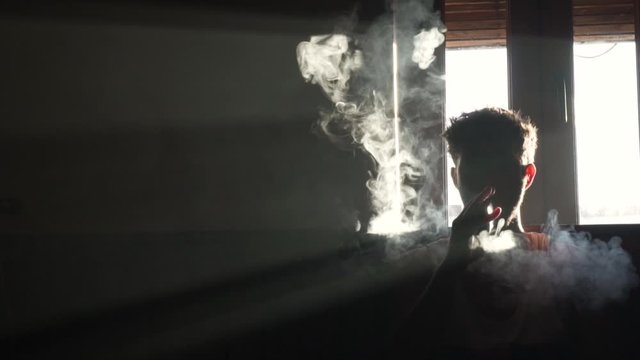 Silhouette of a man vaping in the dark with smoke flowing in slow motion.