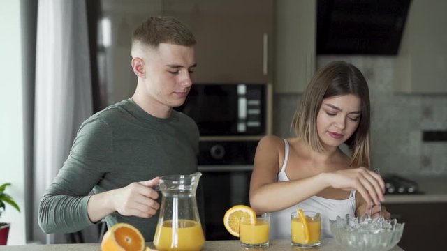 Lovely couple is sitting in a kitchen and drinking an orange juice together. Girl is adding an ice in a glass. Pair are in love and hugging.