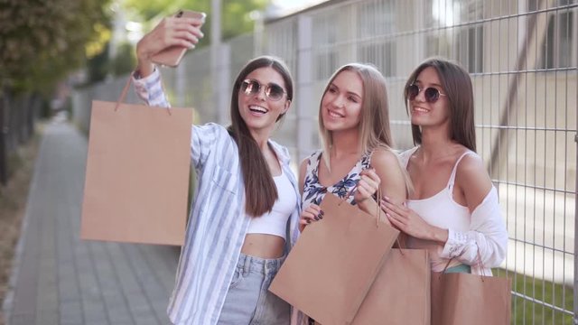 Two beautiful girls are walking on the street and holding a package from shopping centre. They are smiling, making a selfie and posing for a photo.