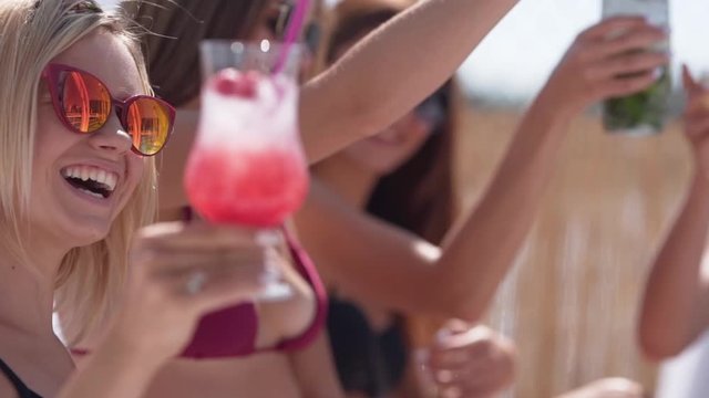 Handheld medium close-up shot of three sexy bikini models dancing and drinking cocktails at the pool