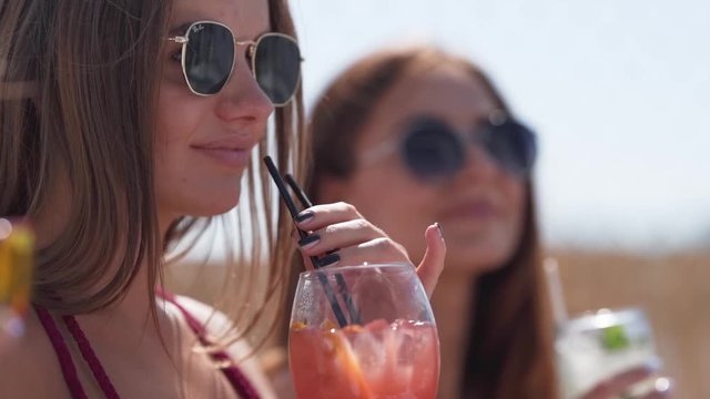 Handheld close-up shot of two sexy beautiful bikini models wearing sunglasses dancing and drinking cocktails at the pool