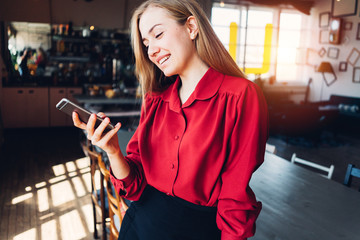 Smiling pretty woman in red and black smart-casual dress holding mobile phone in hands, standing indoors
