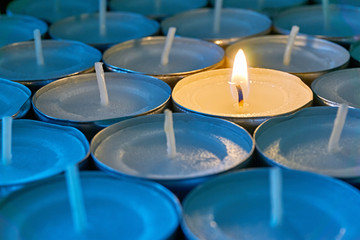 Closeup of tea lights with one being lit. A single warm orange flame between  candles in blue shadow. Christmas, mourning, condolence, memorial, funeral or cremation ceremony concept.   