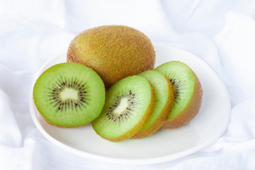 Sliced and whole ripe kiwi fruit in white plate on white tablecloth background.