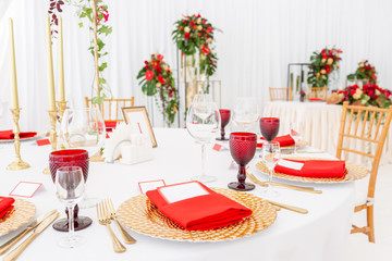 Table setting red napkins and glasses, gold plates. Interior of a wedding tent decoration ready for guests. Decor flowers. Red theme