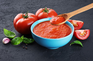 Bowl of tomato soup, tomatoes, garlic and basil on a dark background. Wooden spoon with tomato soup.