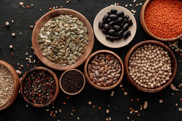 top view of wooden bowls and plates with raw assorted beans, cereals and seeds on dark surface with...