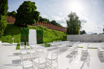 Rows of empty white chairs sitting on a wooden floor. Wedding chairs with flowers at ceremony outdoors