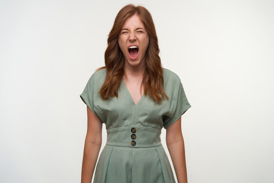 Tired pretty red haired woman posing over white background with wide mouth opened, yawning to camera with closed eyes, wearing vintage dress in pastel color