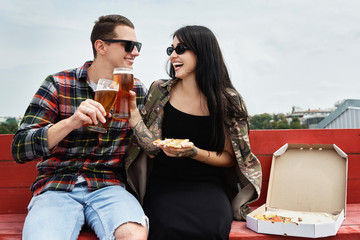 Happy smiling couple eating pizza, toasting and drinking beer at outdoor pub or bar on roof, copy space