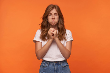 Portrait of unhappy young woman with red curly hair in blue jeans and white t-shirt posing over white background, looking to camera with sad face and raising folded hands