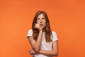 Studio shot of bored beautiful woman in white t-shirt looking upwards, leaning on her chin with drearily face, isolated over orange background