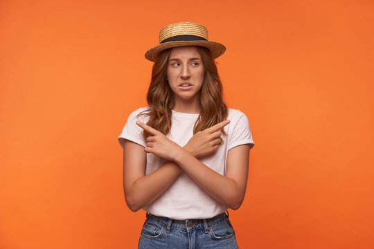 Indoor shot of pretty young redhead curly woman in boat hat standing over orange background with crossed arms on her chast, pointing with index fingers in different directions