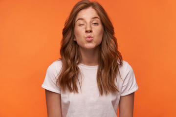 Obraz premium Positive young attractive female posing over orange background with hands down, winking and folding lips in air kiss, wearing casual wear