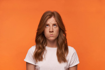 Indoor photo of bewildered redhead curly female looking upwards with confused face, contracting forehead and pouting, posing over orange background