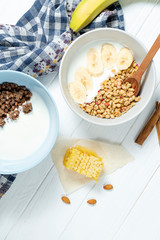 white wheat and chocolate granola with low-fat yogurt in a white bowl in a composition with a spoon, honeycombs, banana, on white wooden background. Healthy breakfast food. Gluten free