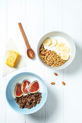 white wheat and chocolate granola with low-fat yogurt in a white bowl in a composition with a spoon, honeycombs, banana fig on white wooden background. Healthy breakfast food. Gluten free