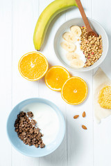 white wheat and chocolate granola with low-fat yogurt in a white bowl in a composition with a spoon, honeycombs, banana, orange on white wooden background. Healthy breakfast food. Gluten free