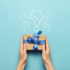 Female hands hold a gift box with a blue ribbon on a blue background with a question mark. Surprise...
