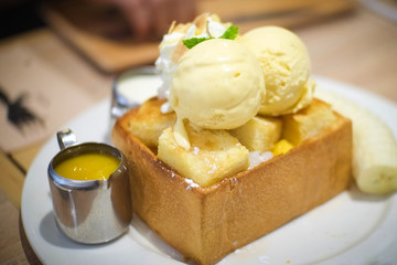 Delicious sweet dessert honey toast with sliced banana mango ice-cream and yellow mango sauce on white plate on wooden table , food dessert happy relaxation concept.