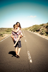 Happy young couple in love walking together on a long straight road - man carry laughing cheerful woman - concept of travel together and have fun