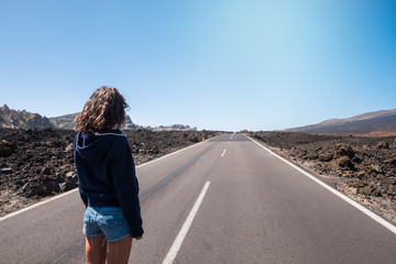 Concept of girls travel alone -standing young caucasian people woman viewed fom back with. jeans shorts looking at the end of a long straight asphalt road at the mountain in desert scenic place