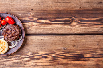 Fresh grilled burger meat cutlets with vegetables - onion rings and cherry tomatoes - on brown vintage plate on wooden background. Grilled food. Copy space.
