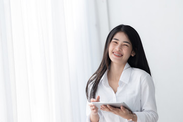 Beautiful Asian woman in white shirt using tablet near window in the morning.