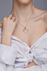 Cropped shot of a lady, wearing white chest tied shirt. She has golden necklace with suspension in a shape of half moon on her neck. Her right hand is touching shoulder. 