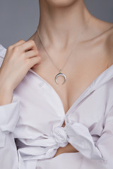 Cropped shot of a lady, wearing white chest tied shirt. She has silver necklace with suspension in a shape of half moon on her neck. Her right hand is touching shoulder. 