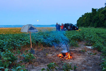 Family traveling and camping, twilight, cooking on the fire. Beautiful nature - field, forest and moon.