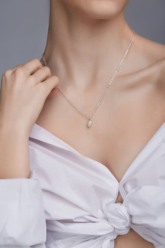 Cropped shot of a lady, wearing white chest tied shirt. She has silver chain with a pendant on her neck. Her right hand is touching shoulder. The photo is taken by grey background. 