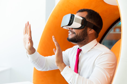 Amazed professional playing VR game in office rest room. Man wearing virtual reality glasses, sitting in interactive chair and touching air with both hands. Simulation concept