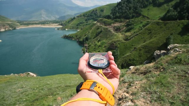The first-person view of a male hand holding a plastic magnetic compass rotates on the side and looking for the right direction in the background of a mountain. The concept of orienteering