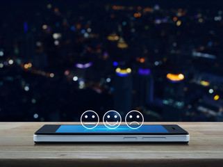 Excellent smiley face rating icon on modern smart mobile phone screen on wooden table over blur colorful night light city tower and skyscraper, Business customer service evaluation and feedback rating