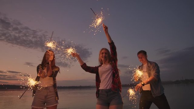 Cheerful male and female friends are running along the beach at sunset, holding sparkling fireworks and runaway lights in slow motion. Dancing and sunset party on the beach.