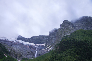Fototapeta premium Beautiful panorama of the Caucasus mountains. Top of mountain range is covered in fog. Clouds lie on mountains. Summer day. Background image for travel and nature.