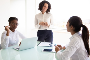 Obraz premium Female business leader explaining project specific to her team. Businesswomen sitting and standing at table, using laptop and talking. Corporate meeting concept