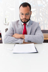 Confident business leader studying contract. Business man wearing suit and eyeglasses, sitting at office table and looking at papers. Paperwork concept