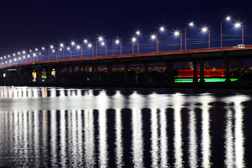 Fototapeta premium The night bridge with beautiful lanterns is reflected in the water on the Dnieper River, in the Dnepropetrovsk city, Ukraine. Evening river landscape in a big city.