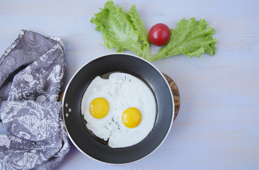  Flat lay fried two eggs with yolk in a pan with a tissue napkin on a blue wooden table with fresh green lettuce and red tomato. Favorite morning breakfast. Top view. With copy space for text.