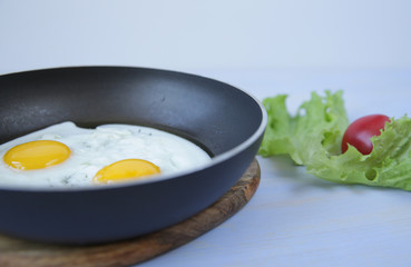 Close-up Fried two eggs with yolk in a frying pan on a blue wooden table with fresh green lettuce and red tomato. Favorite morning breakfast. With copy space for text.