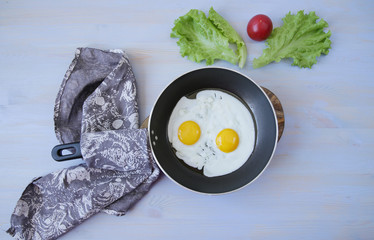  Flat lay fried two eggs with yolk in a pan with a tissue napkin on a blue wooden table with fresh green lettuce and red tomato. Favorite morning breakfast. Top view. With copy space for text.