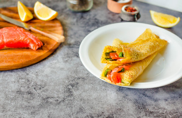 Thin pancakes with red fish on plate, top view.Crepes with filet salmon and cheese, red fish and green avocado on white plate over gray background.  Healthy breakfast