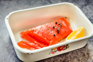 Raw salmon steaks with pepper corns, salt, lemon and olive oil in an enamel bowl, on gray background. Healthy food, diet concept. Preparation for cooking fish with spices