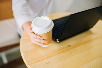 Female hands holding recyclable paper coffee cup at a cafe, working with black laptop. Top view.