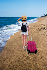 summer vacation and travel concept - back view of young woman tourist in straw hat with suitcase and backpack walking at the beach