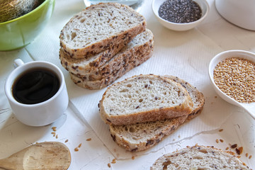 Brown bread with cereals next to a coffee cup on a white wooden table of a rustic kitchen. Seeds and ingredients for home made bakery.