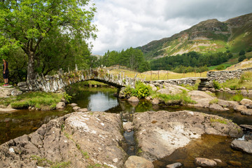 Fototapeta na wymiar Slaters Bridge in the Lake District National Park with fells/mountains in the background and river and rocks in the foreground